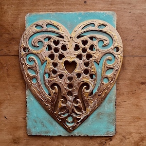 May include: A rectangular plaque featuring a teal-painted wooden base with a distressed finish. Mounted on the front is a metallic gold-toned heart-shaped cutout with intricate, swirling filigree patterns and a small heart-shaped opening.