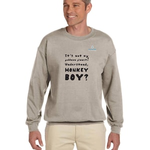 Könnte beinhalten: Ein hellgraues Sweatshirt mit dem Text "It's Not my goddamn planet! Understand, MONKEY BOY?" in Schwarz. Ein kleines blau-weißes Logo befindet sich oben rechts. Die Person trägt eine khakifarbene Hose.