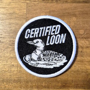 May include: A circular embroidered patch featuring a black background with a white border. The design includes a white illustration of a loon swimming on water, accompanied by the white text "CERTIFIED LOON" positioned above the bird.