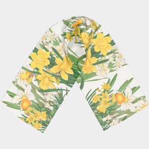 May include: A white scarf with a floral pattern featuring yellow daffodils and white flowers with orange centres. Green leaves and stems are interspersed throughout the design. The scarf is folded in a V-shape.