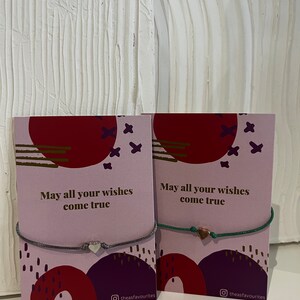Könnte beinhalten: Zwei Armbänder mit Herzanhängern auf rosa und rot gemusterten Karten. Die Karten tragen den Text "May all your wishes come true" und den Instagram-Handle "theasfavourites".