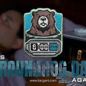 May include: Embroidered patch featuring a brown bear with a blue background and a digital clock displaying 6:00. The text "IT'S GROUNDHOG DAY AGAIN" is below the patch, with the website address www.tacgent.com.