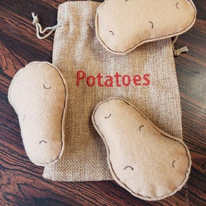 May include: A set of three beige felt potatoes with stitched details and a drawstring hessian bag. The bag has the word "Potatoes" embroidered in red. The potatoes are various sizes and shapes.