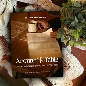 Könnte beinhalten: Ein Buch mit dem Titel Around The Table: 7 Weeks to Knowing and Living Your Christian Faith von Phylicia Masonheimer. Das Cover zeigt ein Foto eines aufgeschlagenen Buches und einer weißen Tasse auf einem Holztisch mit weißem und goldenem Text.