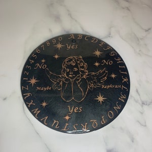 May include: A black circular ouija board with gold lettering and a gold cherub design in the centre. The board features the alphabet, numbers 0-9, "yes", "no", "maybe", and "rephrase".