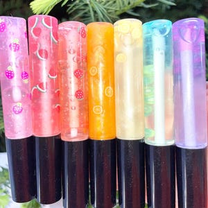 Könnte beinhalten: Sieben Lipgloss-Tuben mit schwarzen Basen. Die Tuben enthalten schimmernde Glosse in Rosa, Orange, Gelb und Blau. Einige Tuben sind mit Fruchtmotiven wie Erdbeeren, Wassermelonen und Zitronenscheiben verziert.
