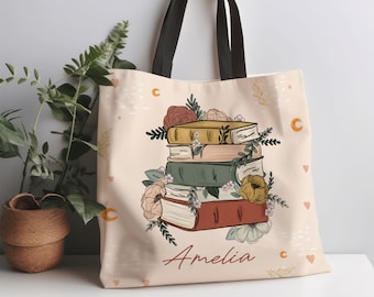 May include: A beige tote bag with black handles featuring a stack of books and floral illustrations. The books are in various colors, including red, green, and yellow, with the name "Amelia" printed on the front. The text "NAME CAN BE CUSTOMIZED" is in the top left corner.