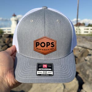 May include: Gray and white trucker hat with a brown leather patch that reads "POPS" above "BRADY & ELLA & THEO." The hat has a curved brim and a mesh back. The Richardson brand label is visible.