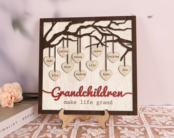 May include: Wooden family tree sign with a dark brown frame and a light wood background. The sign features a tree design with heart-shaped name tags. The words "Grandchildren make life grand" are in red script. The sign is displayed on a wooden easel.