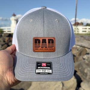 May include: A gray and white trucker hat with a leather patch that says "DAD" with an American flag design. The names "EMMA • ALEX • LUCAS • OLIVIA" are below the word "DAD". The hat has a Richardson 112 Trucker patch.