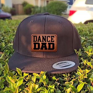 May include: A black snapback hat with a flat brim. The hat features a rectangular patch with the words "DANCE DAD" in bold, black letters on a brown background. The hat is sitting on a bed of green foliage.