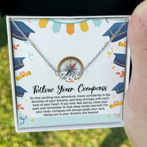 May include: A silver necklace with the name "Zoe" in cursive script, featuring a compass design. The necklace is presented in a gift box with a graduation theme, including mortarboards and the text "Follow Your Compass".