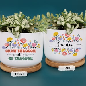 May include: Two white ceramic planters with floral designs and plants. One planter has the text "GROW THROUGH what you GO THROUGH". The other planter has the name "Jennifer". Both planters sit on wooden saucers.