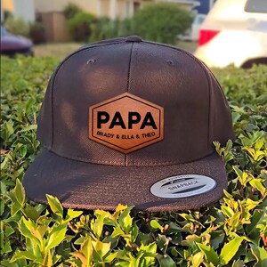 May include: A dark brown snapback hat with a flat brim. The hat features a brown leather patch with the word "PAPA" in bold white letters, and the names "Brady & Ella & Theo" below. The hat is sitting on green foliage.