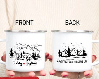 May include: Two white enamel camping mugs with silver rims. One mug features a mountain and tent design with the names "Eddy" and "Sylina." The other mug has a mountain and tree design with the text "Adventure Partners for Life."