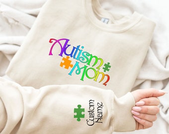 May include: A beige sweatshirt with the words "Autism Mom" in colorful lettering and a puzzle piece design. The sleeve features a green puzzle piece and the words "Custom Name".