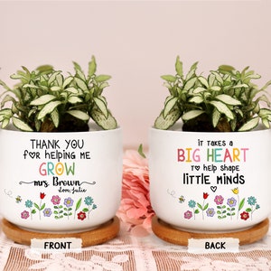 May include: Two white ceramic planters with green plants. One planter says "THANK YOU for helping me GROW" and the other says "It takes a BIG HEART to help shape little minds." Both have colorful floral designs and sit on wooden saucers.