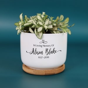 May include: A white ceramic planter with a small green and white plant. The planter has the inscription "In Loving Memory Of Alison Blake 1937-2018" in black script. It sits on a round, natural wood saucer.