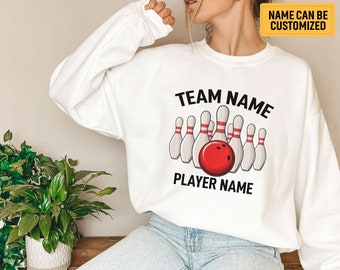 May include: White sweatshirt featuring a bowling design with a red bowling ball and pins. The text "TEAM NAME" and "PLAYER NAME" are printed above and below the bowling graphic. The text "NAME CAN BE CUSTOMIZED" is in a yellow box.