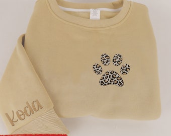 May include: A tan sweatshirt with a leopard print paw design on the front. The sleeve cuff has the name "Koda" embroidered in a matching tan thread. The sweatshirt is folded, showing the paw print and the embroidered name.
