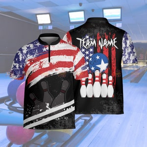 May include: Bowling shirt with an American flag design. The shirt features a distressed look with red and white stripes and blue stars. The back of the shirt has the text "TEAM NAME" and a bowling ball and pins graphic. The shirt has a black collar and a zipper.