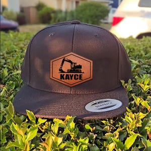 May include: A black snapback hat with a flat brim. The hat features a brown leather patch with an excavator design and the name "KAYCE" in white lettering. The hat is sitting on a bed of green foliage.