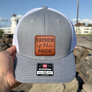 May include: Gray and white trucker hat with a leather patch that reads "FATHER of the BRIDE". The hat has a Richardson 112 adjustable tag.