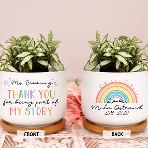 May include: Two white ceramic planters with green and white plants. One planter says "Ms. Browning THANK YOU for being part of MY STORY". The other planter has a rainbow and says "Love, Mila Ostrand 2019-2020". Both sit on wooden saucers.