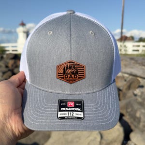 May include: Gray and white trucker hat with a leather patch featuring a bear, trees, and the word "DAD". The hat has a curved brim and a mesh back. The patch is brown and the hat is adjustable.
