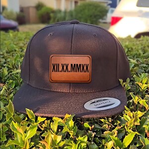 May include: A black snapback hat with a flat brim. A rectangular, brown leather patch with the Roman numerals XII.XX.MMXX is centered on the front. The hat has a snap closure and a white tag that reads "SNAPBACK".