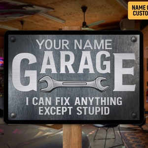 May include: A rectangular metal garage sign with the text "YOUR NAME GARAGE" and "I CAN FIX ANYTHING EXCEPT STUPID" in white. A wrench graphic is centered below the word "GARAGE". The sign is mounted on a wooden post.