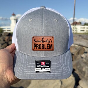 May include: Gray and white trucker hat with a brown leather patch that reads "Somebody's PROBLEM". The hat has a curved brim and a mesh back. The Richardson brand label is visible.