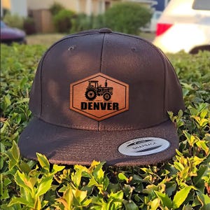 May include: A dark gray snapback cap featuring a hexagonal tan patch on the front. The patch displays a black graphic of a tractor above the text DENVER in bold capital letters. A circular silver and black sticker is visible on the flat brim.