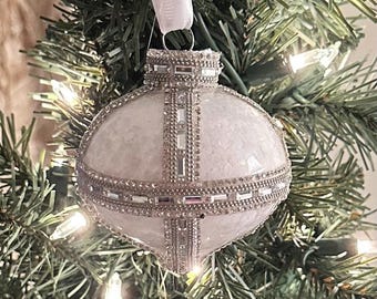 Può includere: Un ornamento natalizio bianco e argento con una superficie strutturata e abbellimenti scintillanti. L'ornamento è rotondo con una fascia argentata e un nastro bianco per appenderlo. L'ornamento è appeso a un albero di Natale verde.
