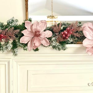 May include: A decorative garland featuring faux evergreen branches, dusted with snow, and adorned with large pink magnolia flowers and rose gold accents. The garland is displayed above a white surface, creating a festive and elegant look.