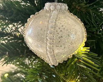 Può includere: Un ornamento natalizio bianco a forma di cuore con un bordo di perline e una finitura scintillante. L'ornamento è appeso a un nastro bianco su un albero di Natale artificiale verde.