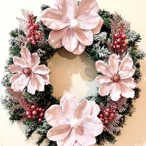May include: A decorative wreath featuring a ring of green pine needles, accented with faux snow. The wreath is adorned with large, light pink flowers and clusters of red berries, creating a festive and elegant look. Perfect for seasonal home decor.