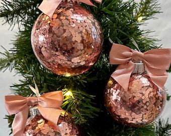 Puede incluir: Tres adornos navideños de purpurina rosa con lazos de satén rosa colgados de un árbol de Navidad verde.