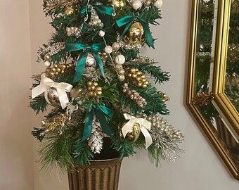 Puede incluir: Un pequeño árbol de Navidad artificial decorado con adornos dorados y blancos, lazos de terciopelo verde y una corona de piña. El árbol está en una maceta dorada y está sentado en una mesa con una flor de terciopelo verde y una flor blanca.