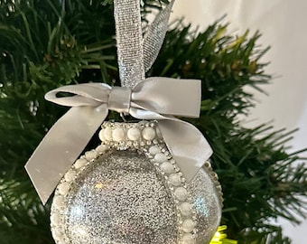 Puede incluir: Adorno navideño plateado con purpurina, ribete perlado y lazo de cinta plateada. El adorno cuelga de una cinta plateada sobre ramas verdes de árbol de Navidad.
