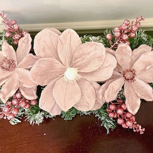May include: A floral arrangement featuring large, light pink magnolia flowers with a central pearl-like accent. The arrangement includes frosted green pine needles, pink berries, and glittery gold leaf accents, creating a festive, decorative piece.