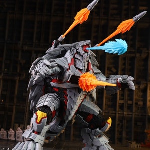 Op de afbeelding: Een grijs en rood robot speelgoed met een dinosaurusachtig ontwerp. De robot heeft meerdere oranje en blauwe projectielwapens. De robot staat op een grijs oppervlak.