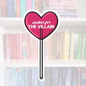 Puede incluir: Una piruleta rosa en forma de corazón con el texto "sucker for THE VILLAIN" en ella. La piruleta está delante de una estantería llena de libros.
