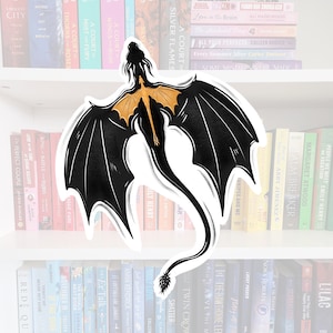 Peut inclure: Un autocollant de dragon noir et or avec des ailes déployées, volant sur un fond blanc. Le dragon a une longue queue et un museau pointu. L'autocollant est sur une étagère avec de nombreux livres.