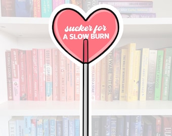 May include: A pink heart-shaped lollipop with the text "sucker for a slow burn" on it. The lollipop is on a white stick with black outlines. The lollipop is in front of a bookshelf filled with books.