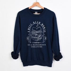 May include: A navy blue sweatshirt with a white graphic design. The graphic features a stack of books with a crescent moon and stars. The text reads "A well-read woman is a dangerous creature."