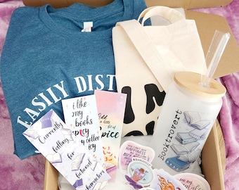 May include: A teal blue t-shirt with the text "EASILY DISTRACTED BY BOOKS" printed on it. The t-shirt is in a cardboard box with a white tote bag, a clear glass tumbler with a bamboo lid and straw, and several bookmarks and stickers with book-themed designs. The stickers have text that reads "You can't buy happiness but you can buy books", "Just one more chapter", "I add a little bit of spice", "Currently faffing for fictional characters", "I like my books spicy & coffee icy", and "Booktrovert".