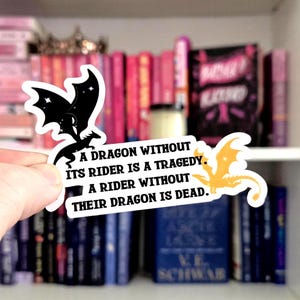 Op de afbeelding: Een witte sticker met zwarte en gouden drakenillustraties en tekst. De tekst luidt: "A DRAGON WITHOUT ITS RIDER IS A TRAGEDY. A RIDER WITHOUT THEIR DRAGON IS DEAD." De sticker wordt voor een boekenplank gehouden.