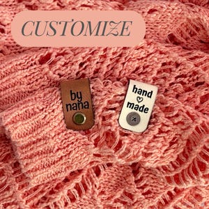 May include: Close-up of a coral-colored knitted fabric with the word "CUSTOMIZE" in a pink oval. Two tags are attached: one brown leather tag with "by nana" and a silver button, and a white tag with "hand made" and a heart.