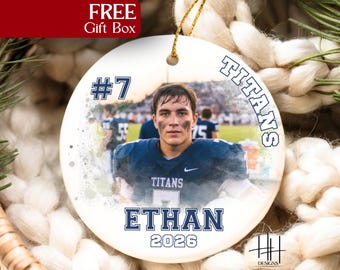 May include: A round white ceramic ornament featuring a photograph of a football player in a dark blue jersey with the text TITANS, the number 7, the name ETHAN, and the year 2026 printed in dark blue block lettering with a gold hanging string.